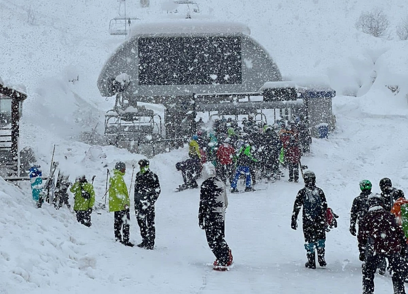 skiers and snowboarders line up for the chairlift, under heavy snow