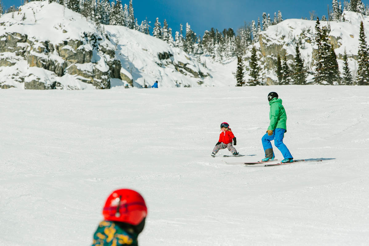 a parent following a tiny child skiing on a wide flat piste, another child's helmet visible in the foreground