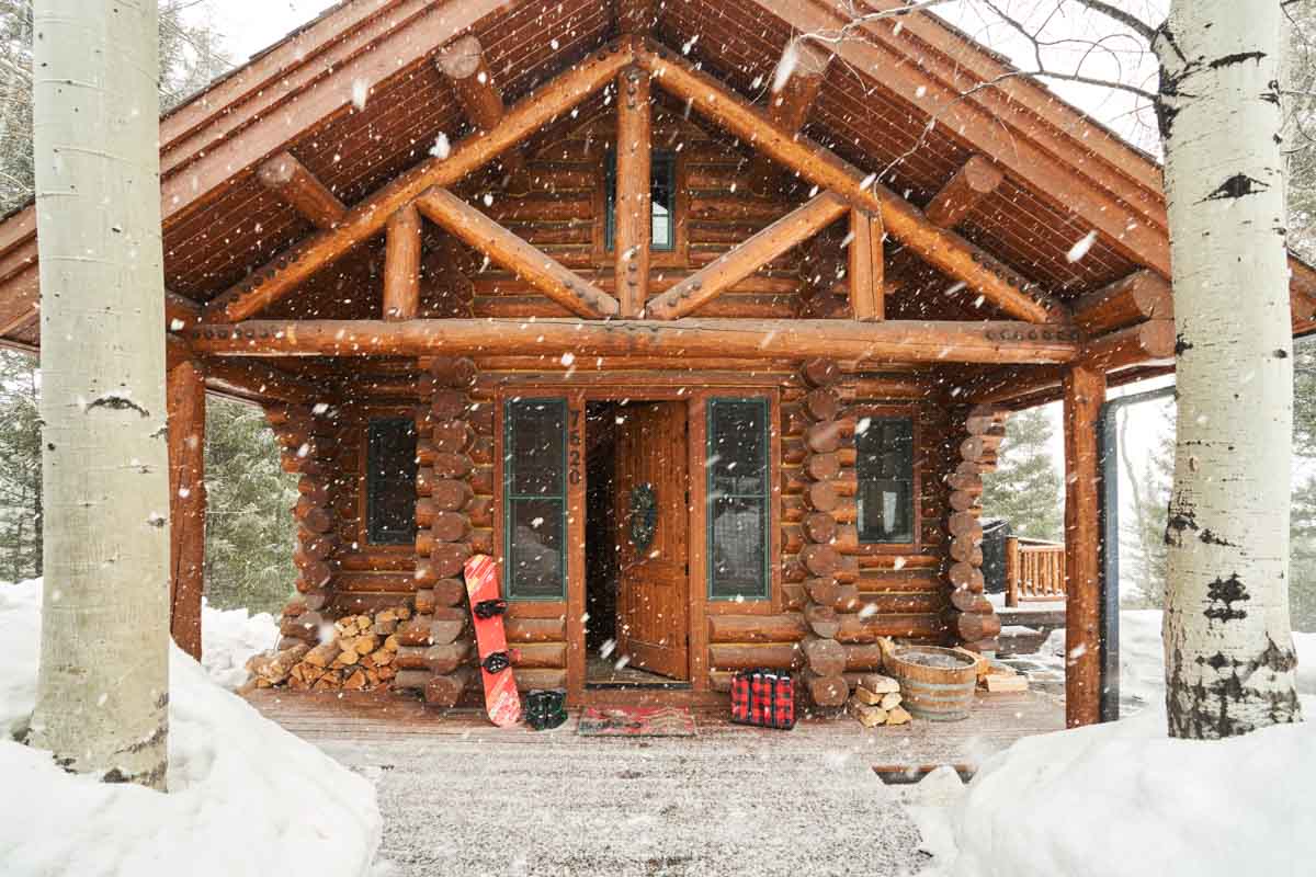 a wood cabin, pictured from the front, snow falling and a snowboard propped up outside