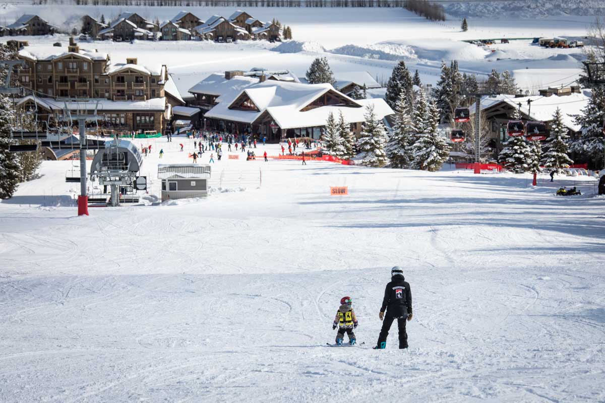 a baby and instructor snowboarders stand on wide, home-run slope of Jackson Hole, the base area in front of them some way
