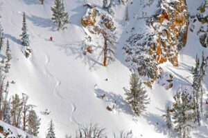 a solo skier in red makes perfect s-shaped tracks off-piste, under cliffs and a few trees. The shot is taken from above