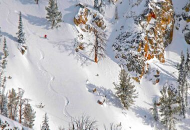 a solo skier in red makes perfect s-shaped tracks off-piste, under cliffs and a few trees. The shot is taken from above