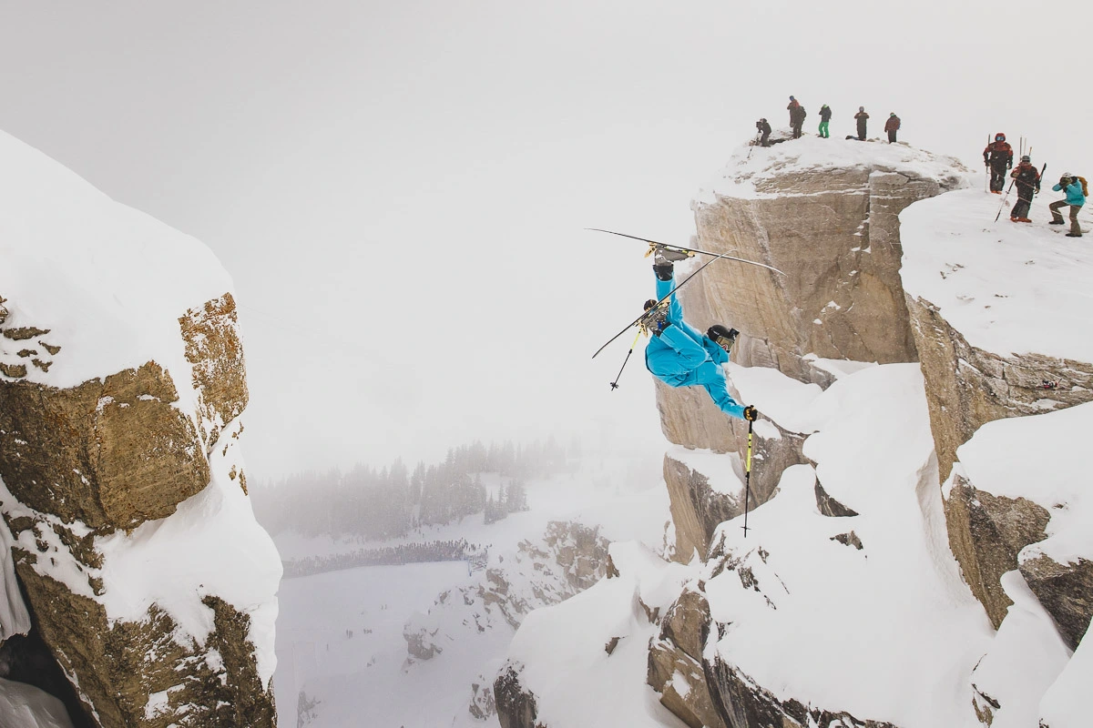 skier launches into couloir, somersaulting with a grab, with skiers along edge of cliff watching