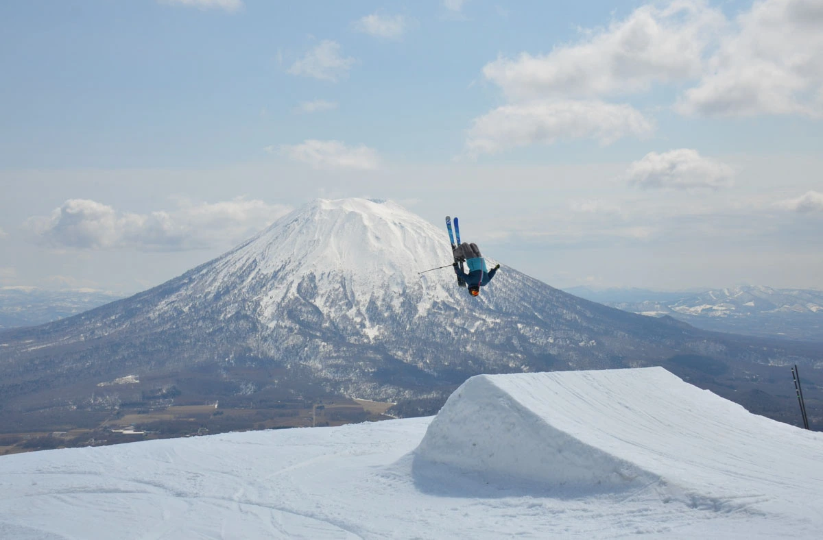 a skier half way round a backflip off a kicker, in front of a snow-topped volcano