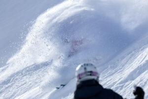 the back of a helmet is in the forefront of a picture, watching a skier pass by almost completely concealed in a spray of snow
