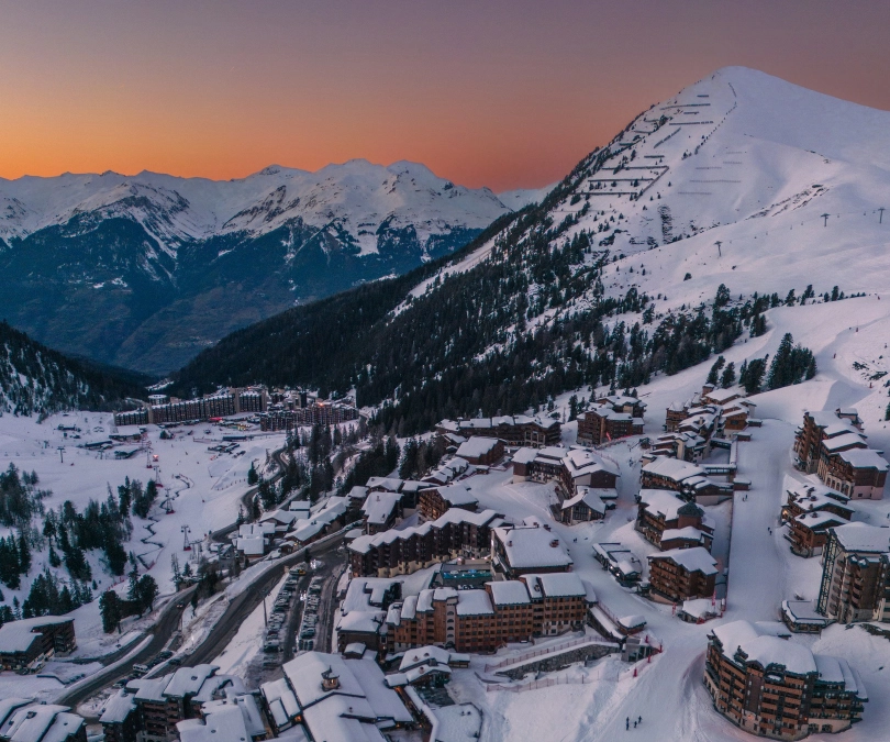 The modern village of La Plagne is photographed, an orange sunset behind the far mountain ridgeline, snow on roofs in near-ground