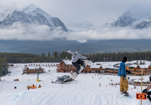 a snowboarder takes air on a jump in the park, above a base lodge and high mountains in the background