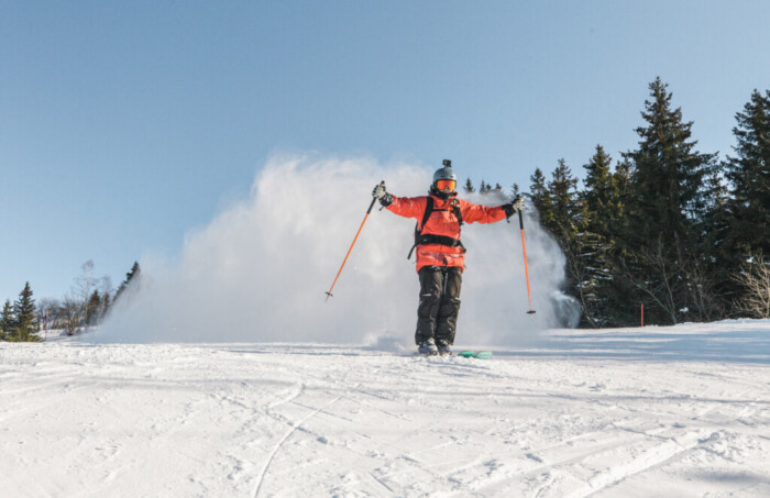 a cloud of dry powdery snow followd a skier in red jacket, arms out, skis close together, on a mellow piste on a sunny day