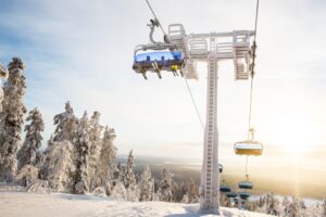 three skiers on a chairlift, bubble closed, ride upwards, in the very snowy Levi ski resort with snow-laden trees.