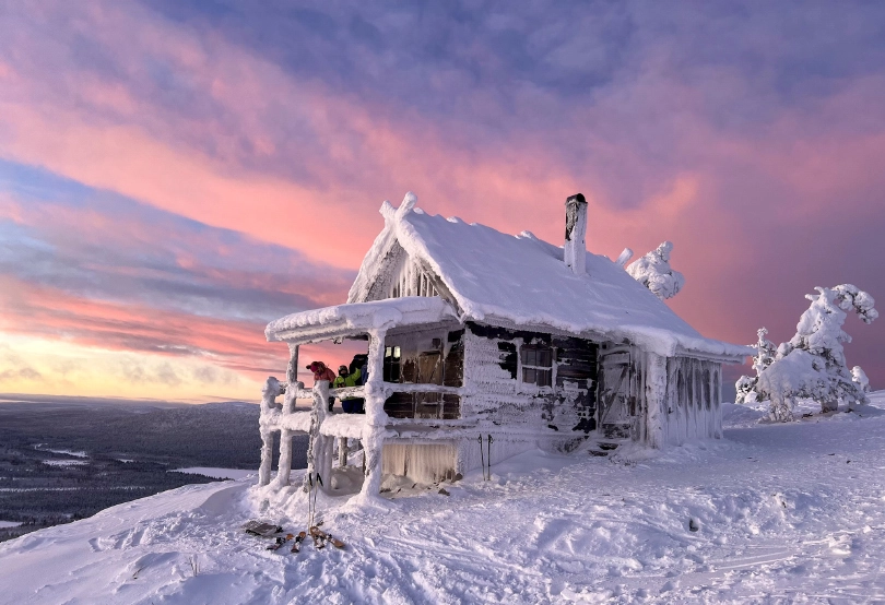 A ski cabin, covered in snow and rime ice, pictured at sunset with a pastel pink and blue sky