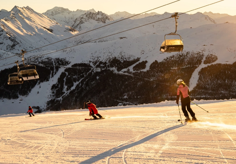 three skiers lazily ski down corduroy piste with low, orangey light shone by the late-afternoon sun (no doubt)