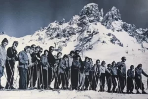 a lineup of skiers atop a mountain, in a vintage photo