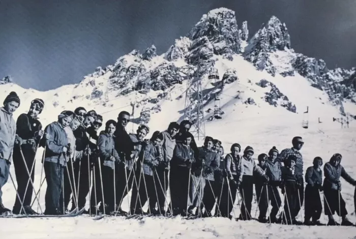 a lineup of skiers atop a mountain, in a vintage photo