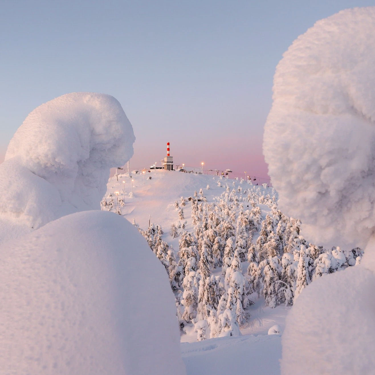 snow monsters in the foreground frame a red and white striped tower at the top of a chairlift on the next mountain top, with a pastel pink sky