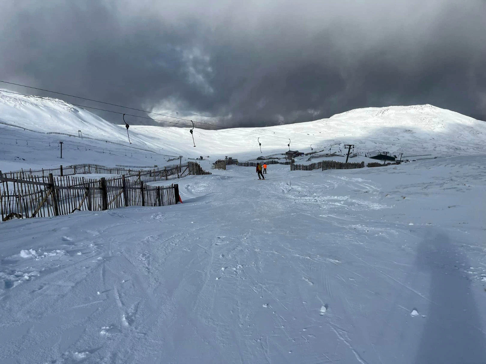 A Scottish ski hill with snow fences surround a thin piste on a grey day, with two skiers skiing