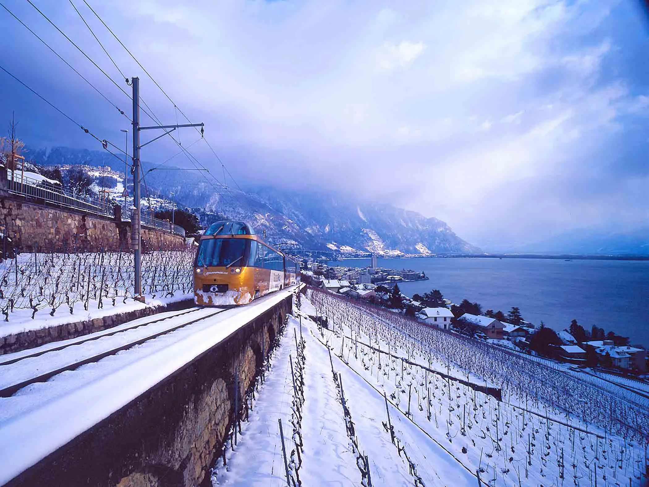 a train travels along snowy tracks by a lake, thorugh a vineyard