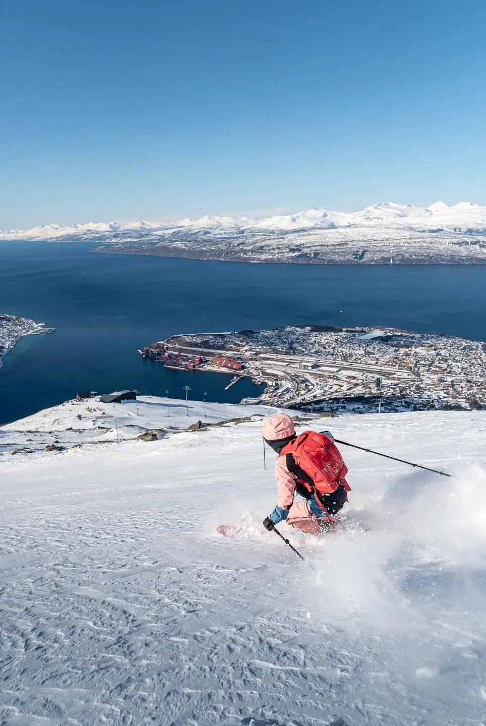 a skier in pink, with a red pack, skis down a smooth-ish, slightly wind affected un-pisted run, above a blue-water fjord, leaving no doubt it's in Norway