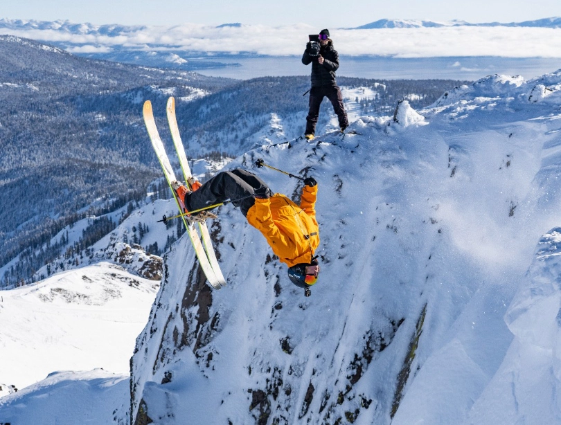 a skier mid-way through a backflip into a couloir, a photographer standing on the rock behind