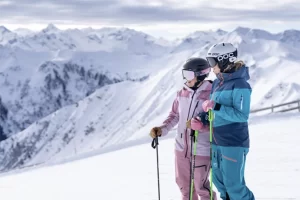 two female skiers in helmets and colourful ski outfits stand side by side on the slopes, looking ahead
