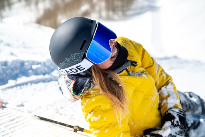 a woman in yellow ski jacket, lies on the snow, smiling, ski goggles and helmet covering most of her face