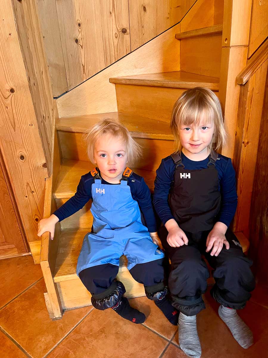 two young children sit on stairs of wooden-clad chalet wearing salopettes, on their family ski holiday to 3 Valleys