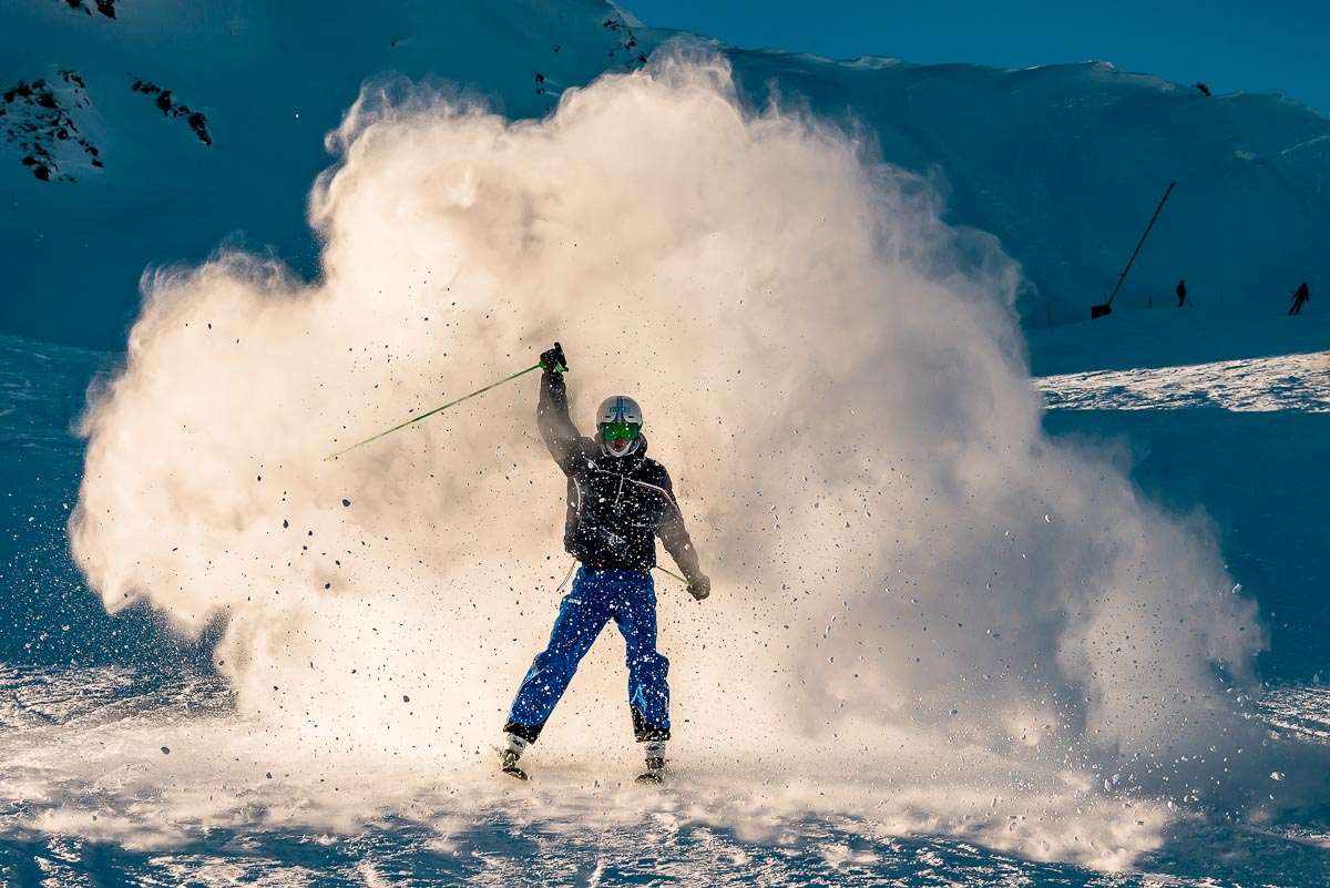 skier raises one arm in celebration, skiing towards camera through a cloud of powdery snow smoke