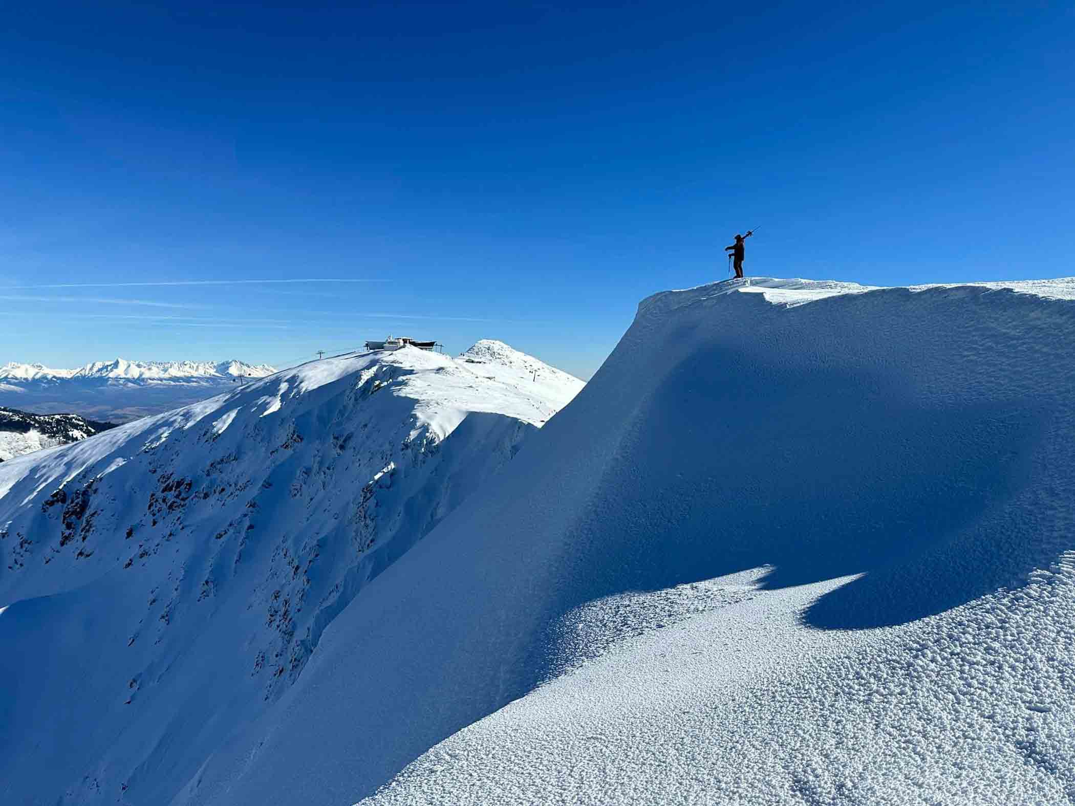 a skier, skis on shoulder, stands on a cornice overlooking a snowy slope