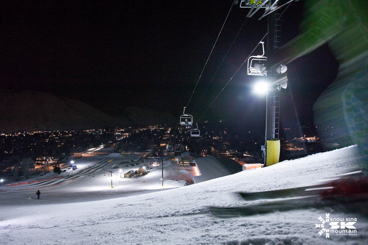 a blurry skier is just in shot of a ski slope at night, the town's lights alight below