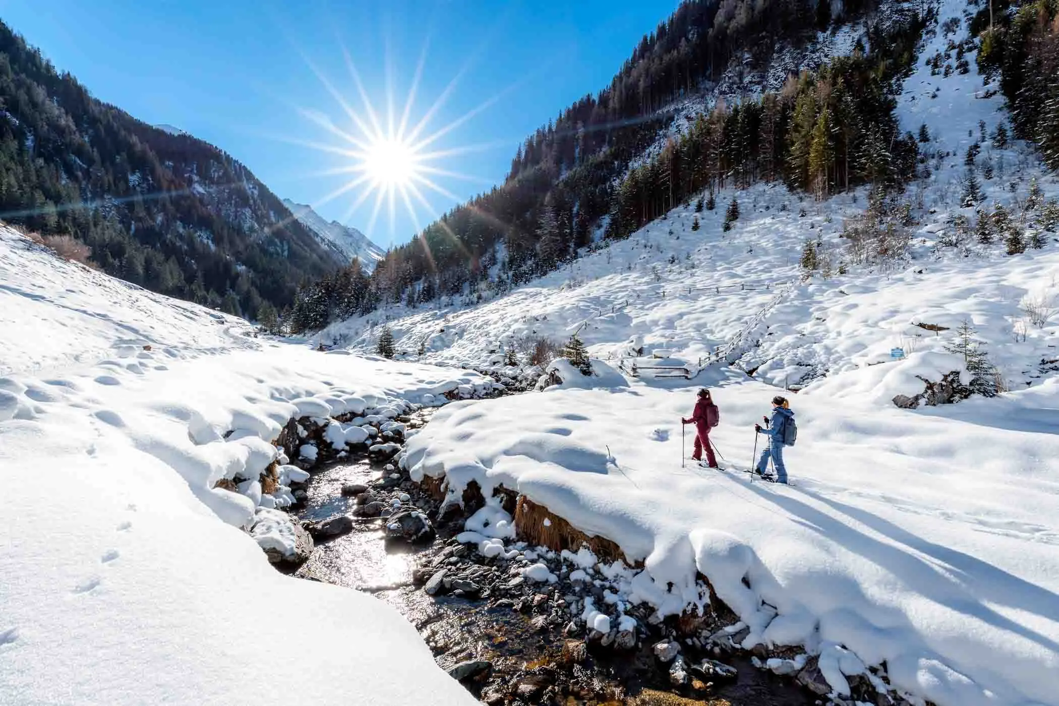 two snowshoers walk through a snowy valley next to a river