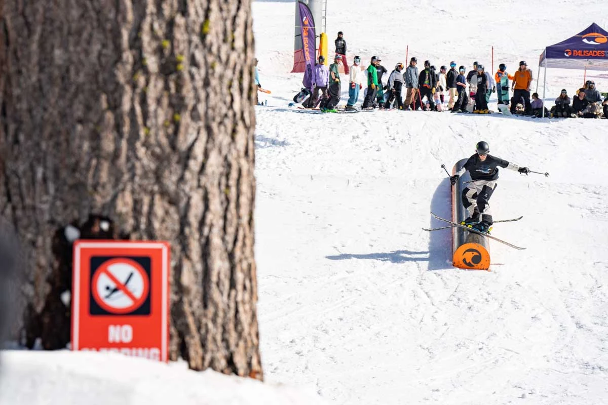 a skier slides a tube-box in the snow park, a crowd behind and a blurred out warning sign of a stick man sledging face-forwards nailed to the tree in the foreground