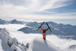 Man, skis in hands raised above head, stands on a snow-lip at the top of a mountain with a huge vista behind