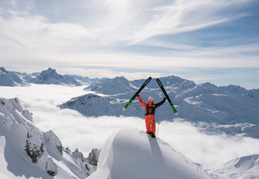 Man, skis in hands raised above head, stands on a snow-lip at the top of a mountain with a huge vista behind