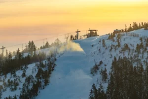 a yellow-orange sunset makes a lovely backdrop for a chairlift top station, a snow machine pumping out snow in the foreground