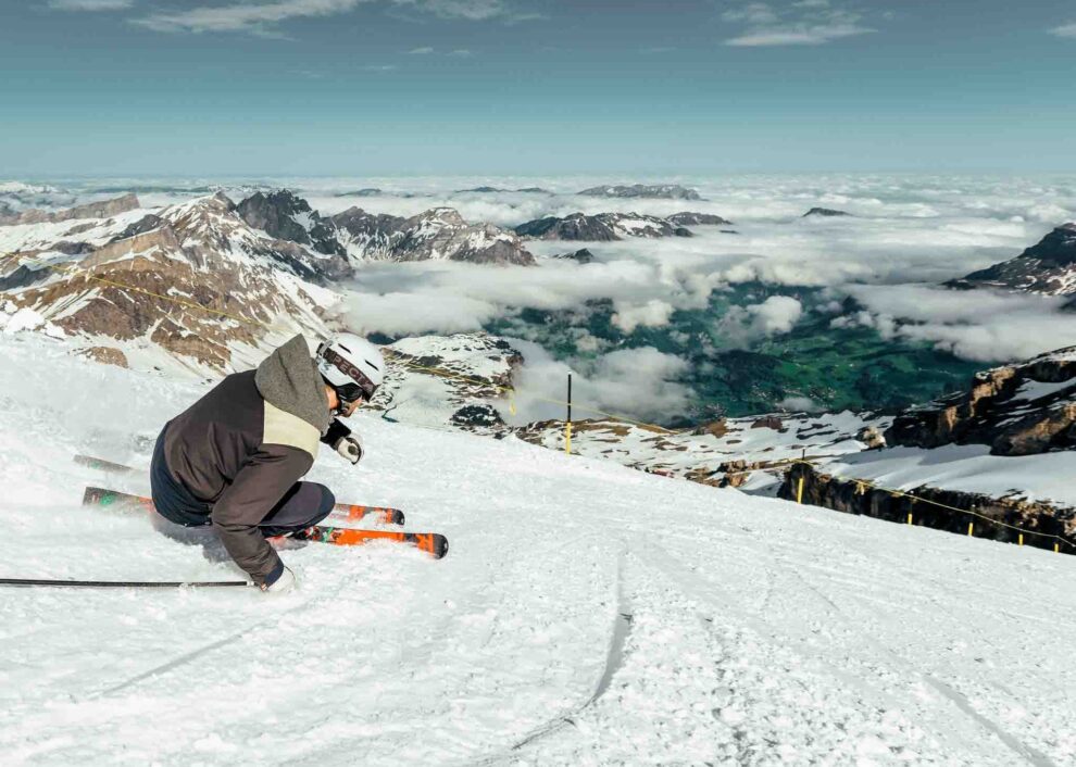 a skier carves deep on spring like snow on-piste, the green valley below almost entirely covered in cloud with mountain peaks poking through