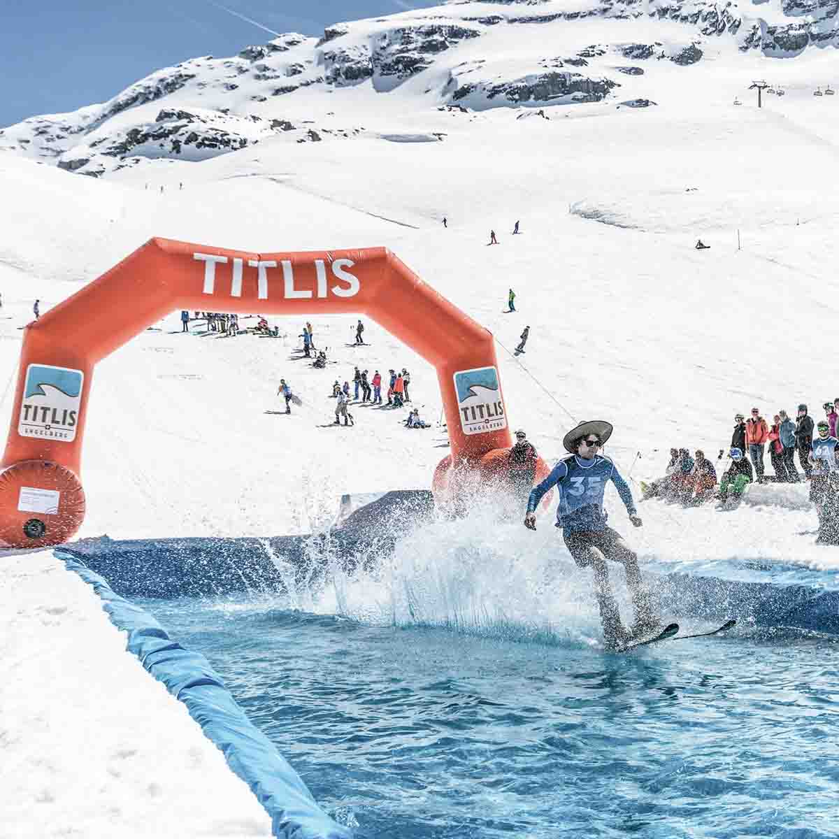 water skimming shenanigans at the top of a ski area, surrounded by white piste and spectators as a man in shorts and a panama hat skims across water on skis