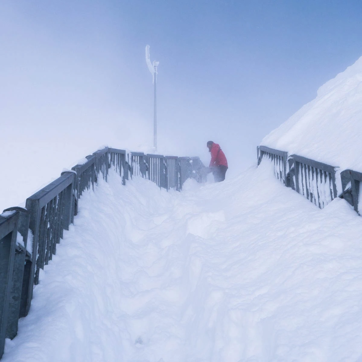 hip-high powdery snow has blown into a walkway, a man in red at the top digging it out under a blue sky