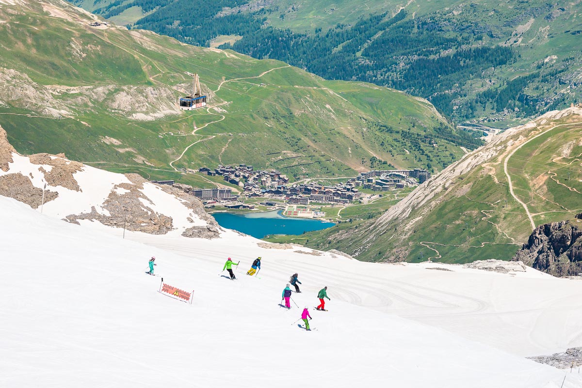 a green valley and blue lake down below is contrasted against a ski slope with a group of skiers on the piste, skiing a glacier during summer