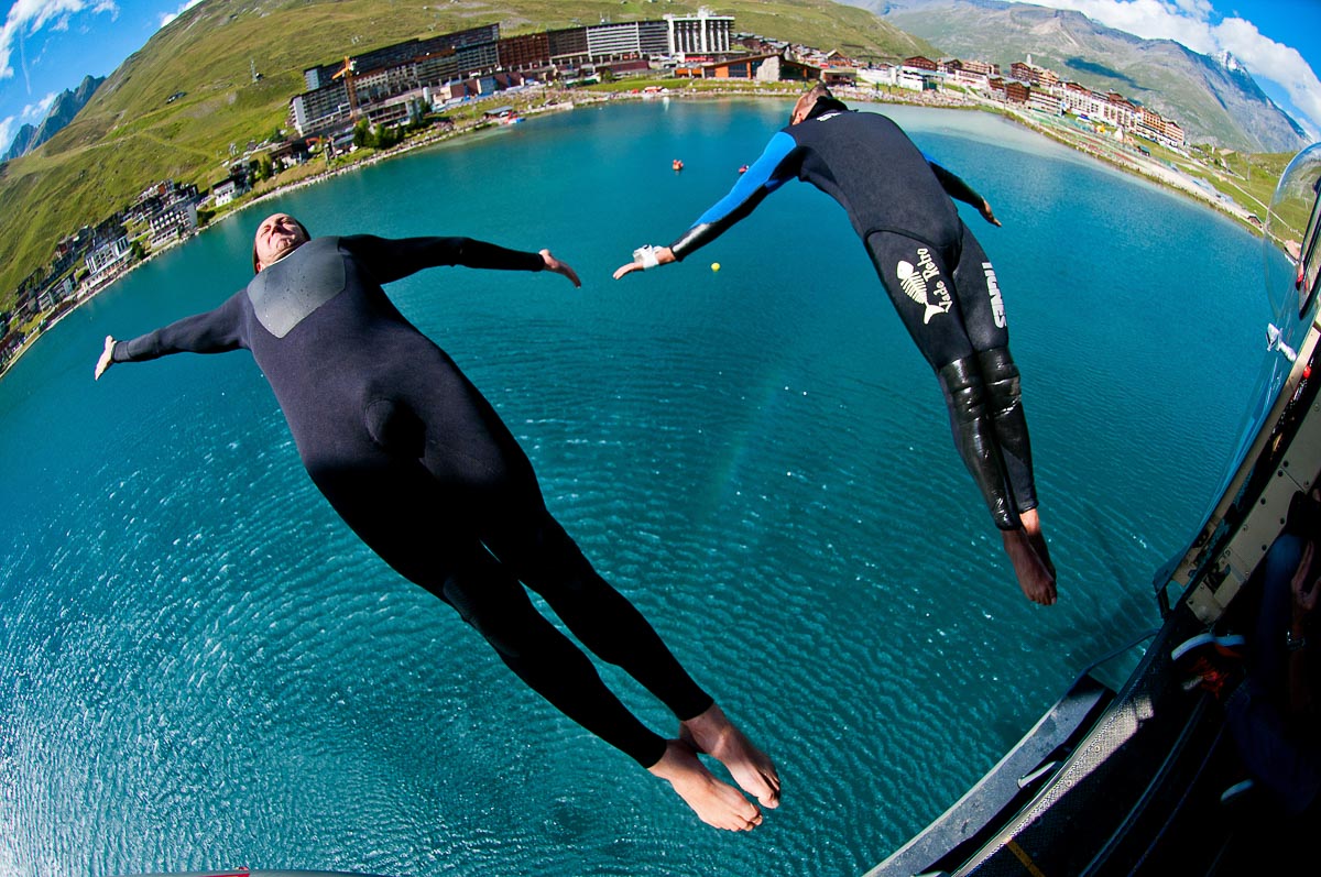 two people dive backwards out of a helicopter into an alpine lake, wearing wetsuits