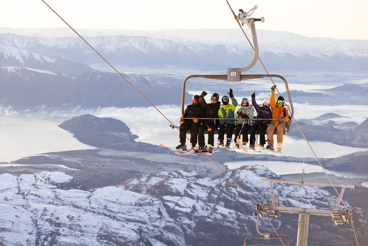 six sit on a chairlift with arms in the air to camera - the lakes and snowy fields of the Wanaka low-lands gleaming in the far distance beneath them