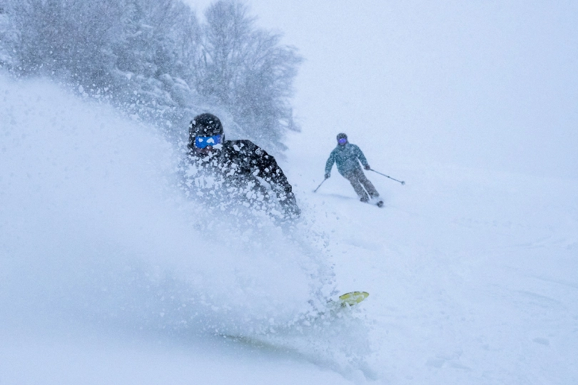 A snowboarder and skier, ride while its snowing heavily