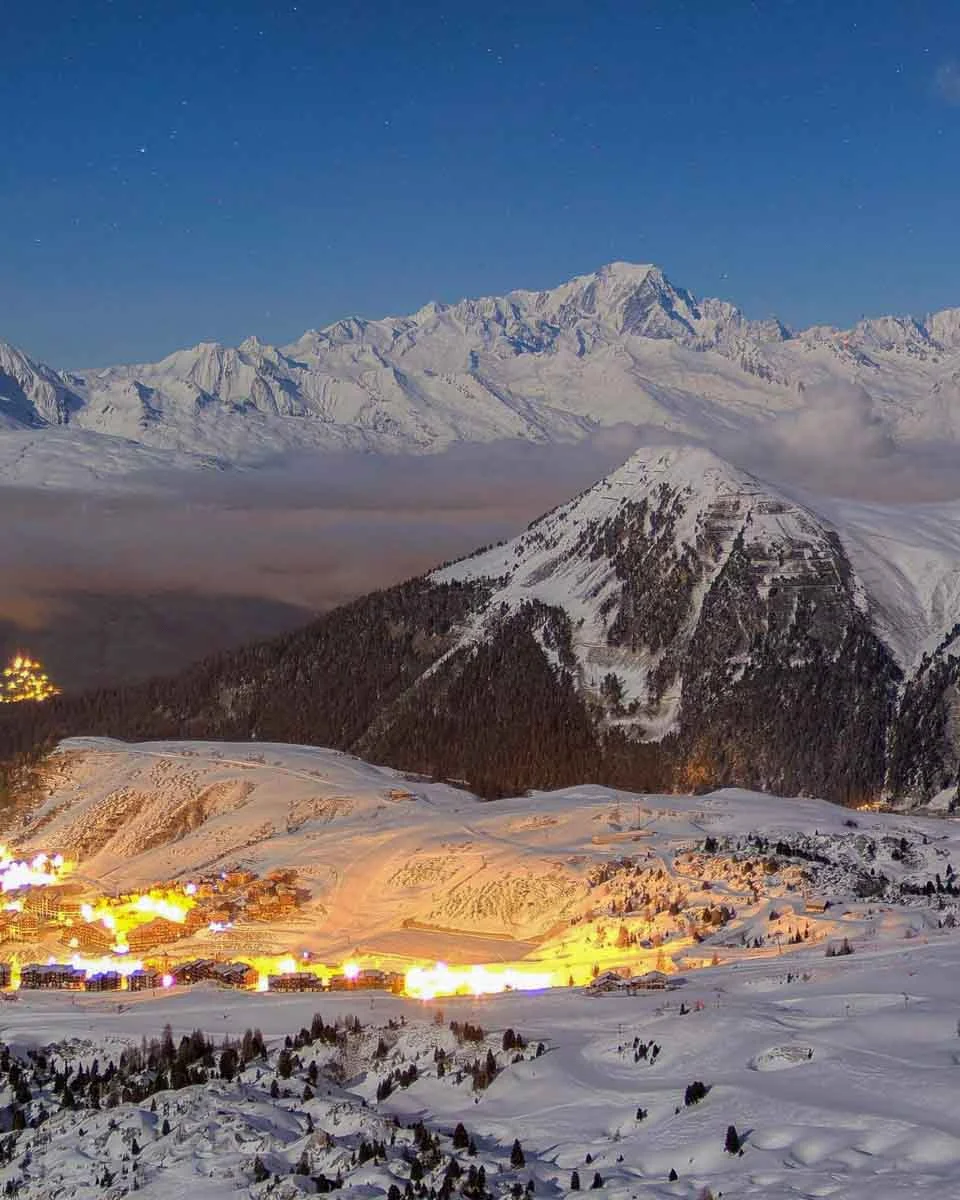A huge, snowy mountain massif is white and looks majestic in the background, behind a cloud-filled valley and lit-up village in the near-ground