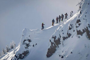 a line of half-a-dozen or so skiers standing on a ridge, steep drop ins to the visible side