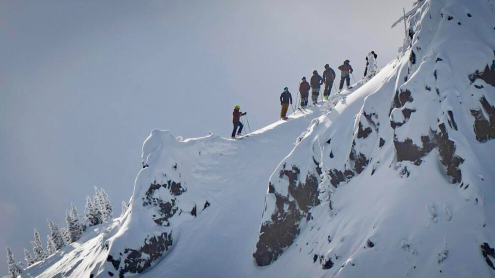 a line of half-a-dozen or so skiers standing on a ridge, steep drop ins to the visible side