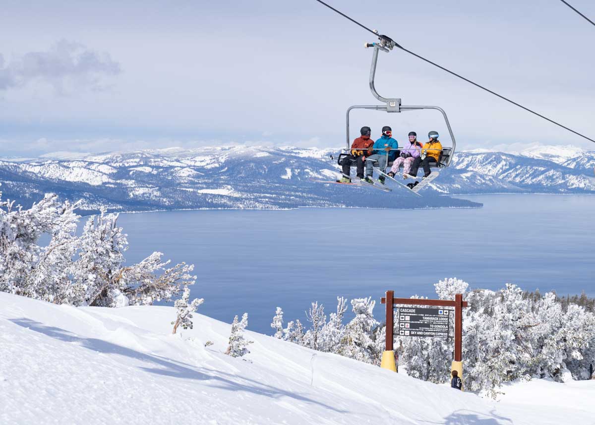 four in colourful ski gear sit on a chairlift, a lake sits behind them, below the snowy hills