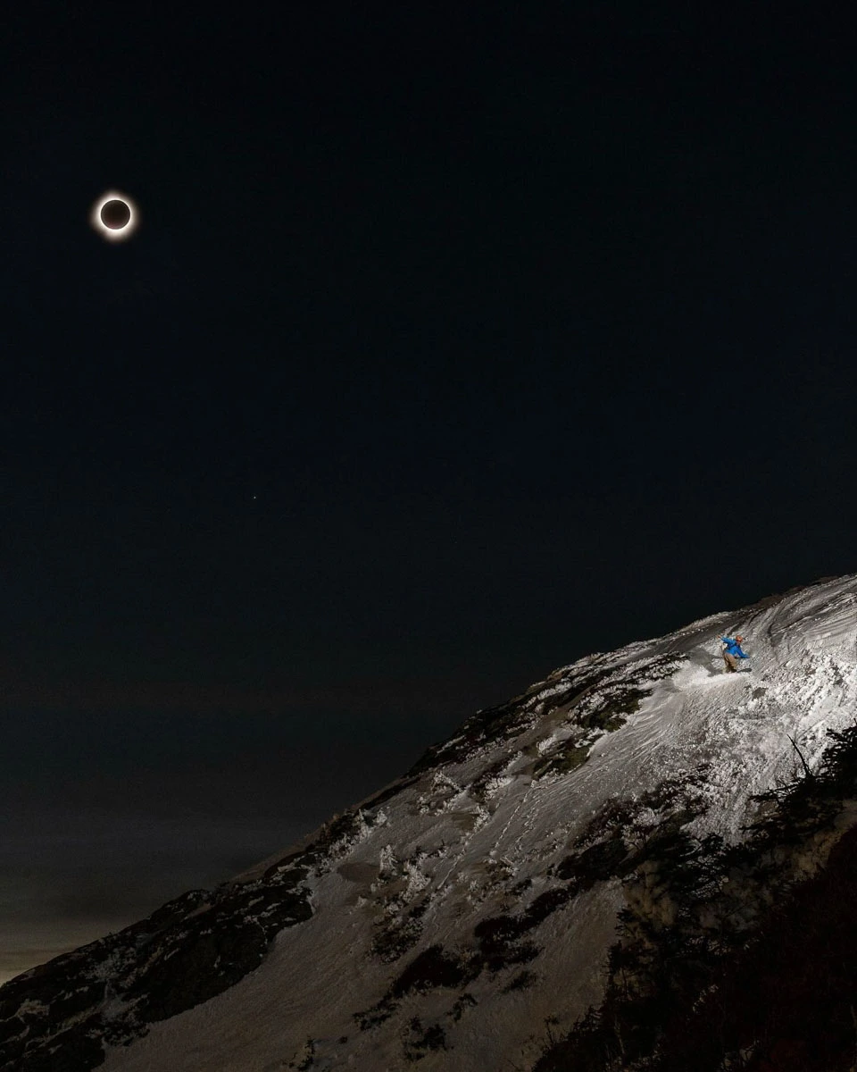 the moon covers the sun, in an eclipse shot, with a snowy mountain section illuminated with a skier just visible