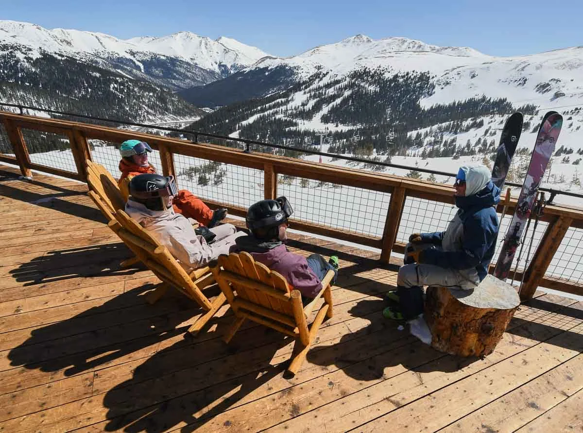 a group of four sit on a wooden terrace, chilling in the sun bu the ski slopes