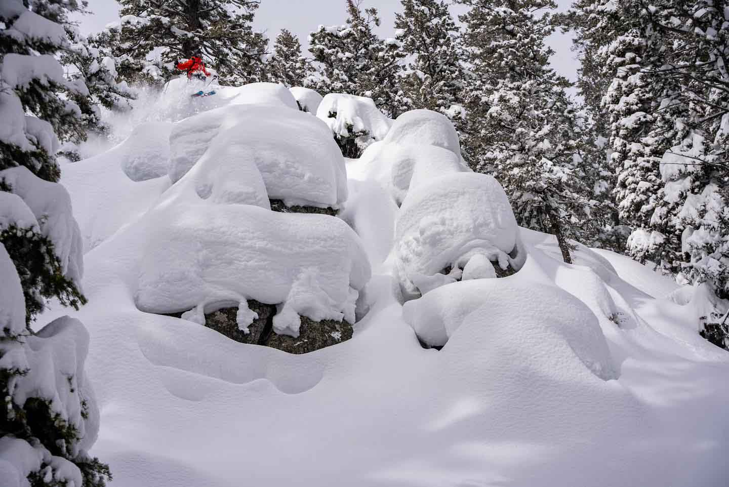 huge snow depth shown piled up on a cliffy rock section, with a skier in red, tiny, against the big drops they're making
