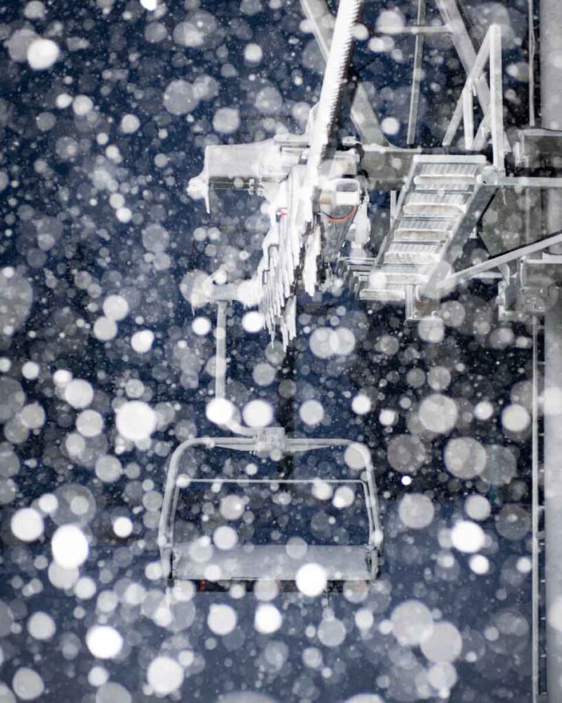 snow flakes out-of-focus dominate the image, a chairlift at night the background