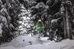 a skier in green takes air popping off a natural snow jump in the big, snowy fir trees that have got to be in North America