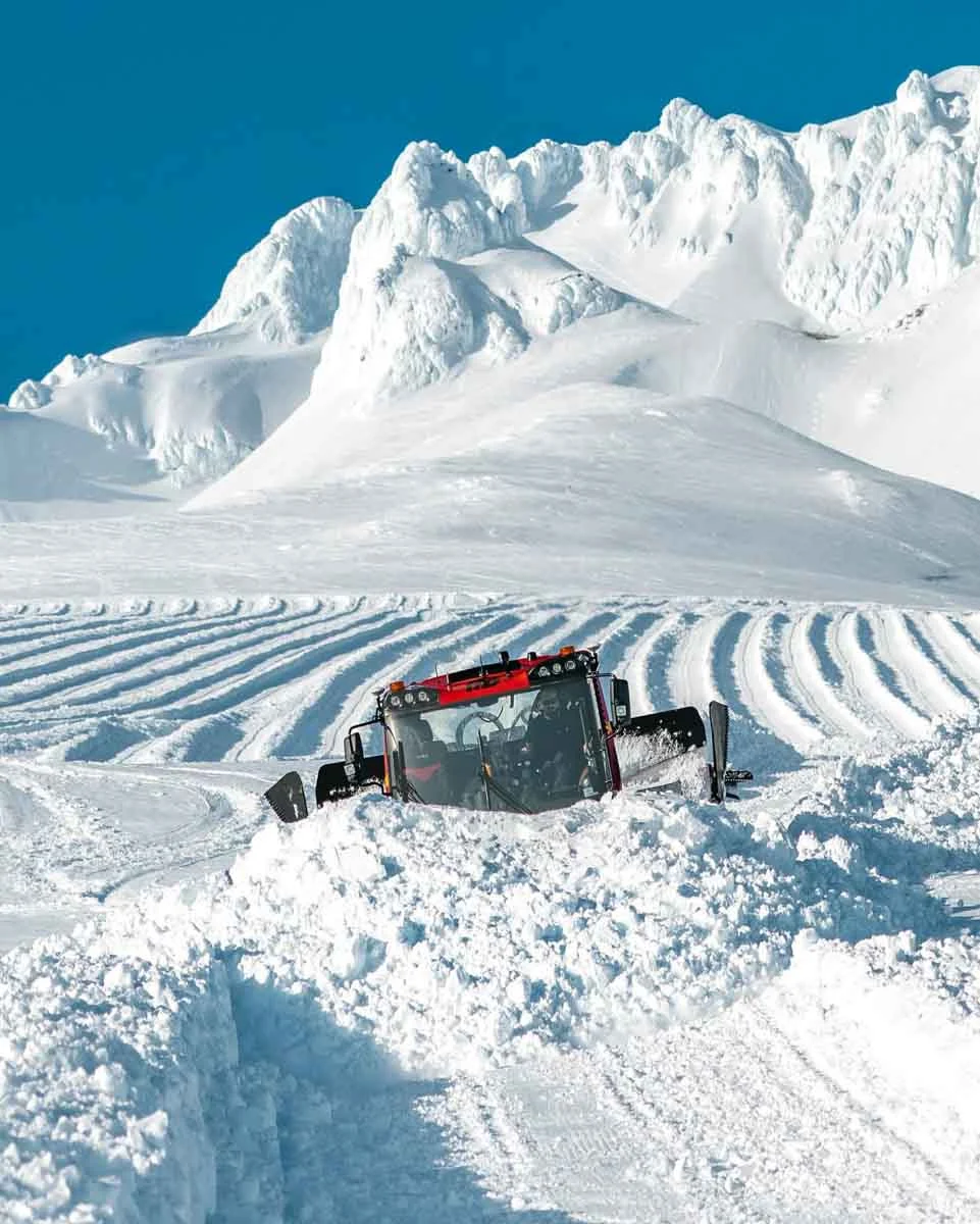 a red piste basher struggles pushing huge pile of snow, in front of a snow farm in the high, very snowy mountains